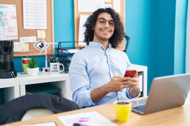Genç Latin iş adamı ofiste dizüstü bilgisayar ve akıllı telefon kullanıyor.