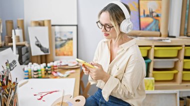 Sanat stüdyosunda akıllı telefon ve kulaklık kullanarak masada oturan genç sarışın kadın sanatçı.