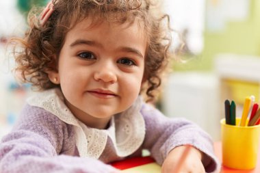 Adorable hispanic girl preschool student sitting on table drawing on paper at kindergarten