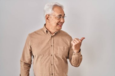 Hispanic senior man wearing glasses smiling with happy face looking and pointing to the side with thumb up. 