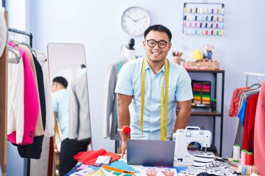Young chinese man tailor smiling confident using laptop at tailor shop