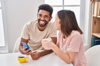 Man and woman couple sitting on table drinking coffee using smartphone at home