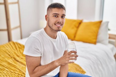 Young hispanic man drinking glass of water sitting on bed at bedroom