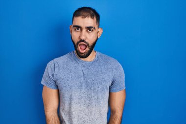 Mavi arka planda duran sakallı bir Orta Doğulu. Şok suratlı. Şüpheci ve alaycı görünüyor. 