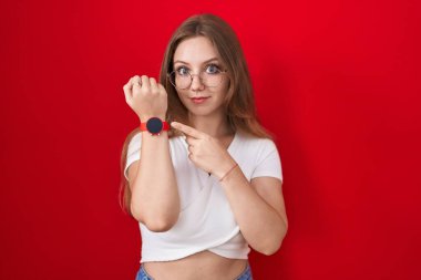 Genç beyaz kadın kırmızı arka planda dikiliyor. Aceleyle zamanı, sabırsızlığı ve rahat bir ifadeyle kameraya bakıyor. 