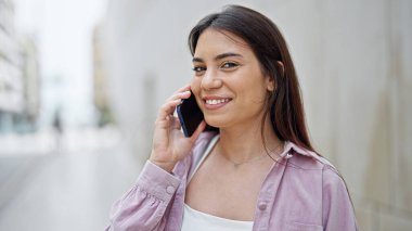 Genç, güzel, İspanyol bir kadın gülümsüyor ve caddede akıllı telefondan konuşuyor.
