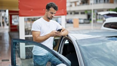 Young hispanic man using smartphone leaning on car at street