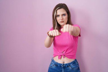 Sarışın, beyaz bir kadın pembe arka planda duruyor. Yumruklarını yumrukluyor, saldırıyor, saldırıyor, tehdit ediyor ve şiddet uyguluyor. 