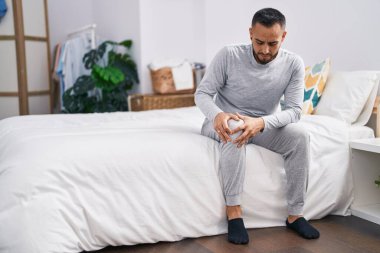 Young hispanic man suffering for knee injury sitting on bed at bedroom