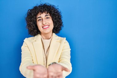 Mavi arka planda kıvırcık saçlı, elleri avuçlarında gülümseyen ve el hareketi yapan esmer bir kadın. Bekleme ve koruma 