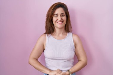 Pembe arka planda duran esmer kadın ellerini kavuşturup gülümseyerek gevşemiş ve neşeli gülümsüyor. Başarı ve iyimserlik 
