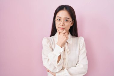 Pembe arka planda duran genç Çinli kadın kameraya kollarını kavuşturup gülümseyerek ve çenesini kaldırarak bakıyor. Olumlu düşünmek. 