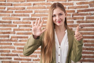 Kafkasyalı genç bir kadın duvarın arkasında duruyor. Arka planda 6 numaralı parmaklarıyla işaret ediyor. Kendinden emin ve mutlu bir şekilde gülümsüyor.. 