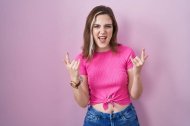 Blonde caucasian woman standing over pink background shouting with crazy expression doing rock symbol with hands up. music star. heavy music concept. 