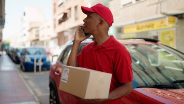 Genç latin teslimat işçisi elinde paketle caddede akıllı telefondan konuşuyor.