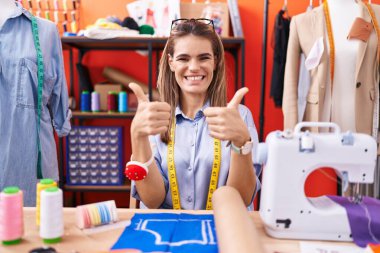 Atölye başarı tabelasındaki İspanyol kadın terzi el hareketleriyle olumlu jest yapıyor, gülümseyerek ve mutlu. Neşeli ifade ve kazanan jest. 