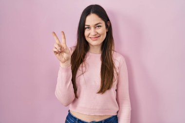 Genç esmer kadın pembe arka planda duruyor. İki numarayı gösterip işaret ediyor. Kendinden emin ve mutlu bir şekilde gülümsüyor.. 