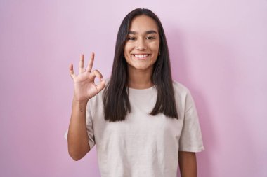 Pembe arka planda duran genç İspanyol kadın gülümsüyor ve el ve parmaklarıyla 