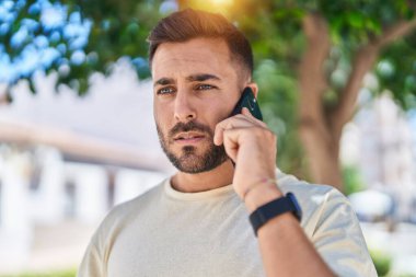 Young hispanic man talking on smartphone with serious expression at park