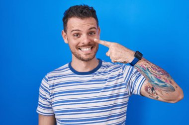 Young hispanic man standing over blue background pointing with hand finger to face and nose, smiling cheerful. beauty concept 