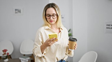 Genç sarışın iş kadını akıllı telefon kullanıyor. Ofiste kahve içiyor.