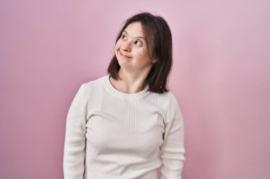 Down sendromlu bir kadın pembe arka planda durup gülümsüyor ve yan tarafa bakıyor.. 