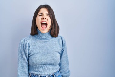 Young hispanic woman standing over blue background angry and mad screaming frustrated and furious, shouting with anger. rage and aggressive concept. 