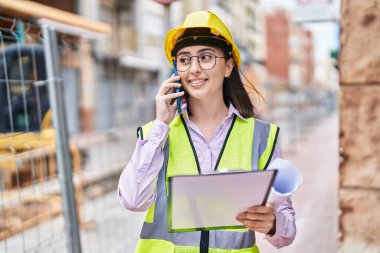 Young hispanic woman architect talking on the smartphone reading document at street