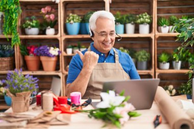 Çiçekçide çalışan, gri saçlı orta yaşlı bir adam. Video çekiyor. Başparmağıyla yan tarafa işaret ediyor. 