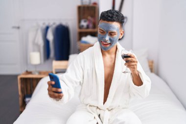 Young latin man sitting on bed make selfie by smartphone drinking wine at bedroom