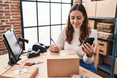 Genç ve güzel İspanyol kadın ecommerce iş işçisi. Ofisteki pakette akıllı telefon yazısını kullanıyor.