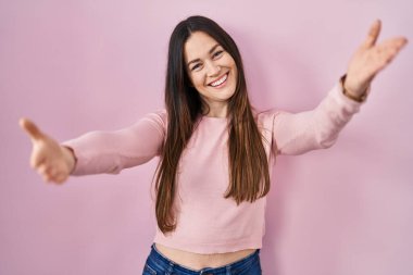 Young brunette woman standing over pink background looking at the camera smiling with open arms for hug. cheerful expression embracing happiness. 