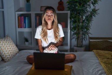 Genç sarışın kadın evde yatağında oturmuş dizüstü bilgisayardan film seyrediyor. Kameraya bakıyor ve öpüşüp öpüşüyor. Sevimli ve seksi. aşk ifadesi. 