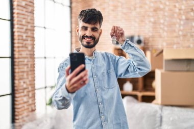 Genç İspanyol adam yeni evinde elinde anahtarla akıllı telefondan selfie çekiyor.
