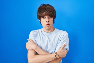 Hispanic young man standing over blue background shaking and freezing for winter cold with sad and shock expression on face 