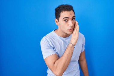 Latin asıllı genç bir adam mavi arka planda el ele tutuşup gizli söylentiler yayıyor, kötü niyetli konuşmalar fısıldıyor. 