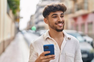 Genç Arap adam sokakta akıllı telefon kullanarak kendinden emin gülümsüyor.