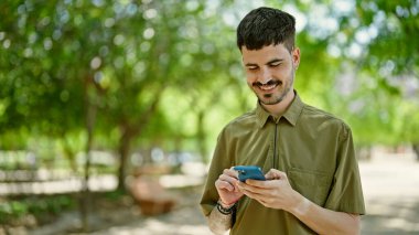 Genç İspanyol adam parkta gülümseyen akıllı telefon kullanıyor.