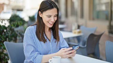 Akıllı telefon kullanan genç ve güzel bir İspanyol kadın kahve dükkanının terasında oturmuş gülümsüyor.