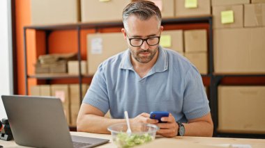 Ofisteki masada oturan dizüstü bilgisayar ve akıllı telefon kullanan gri saçlı bir adam.
