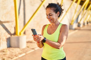 Middle age hispanic woman working out with smartphone at promenade