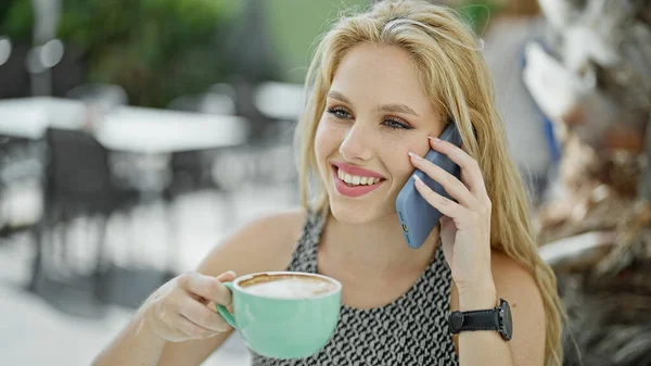 Young blonde woman drinking cup of coffee talking on smartphone at coffee shop terrace