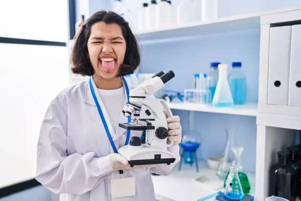 Młoda Latynoska Kobieta Pracująca Laboratorium Naukowca Trzymająca Mikroskop Wystający Języka — Zdjęcie stockowe
