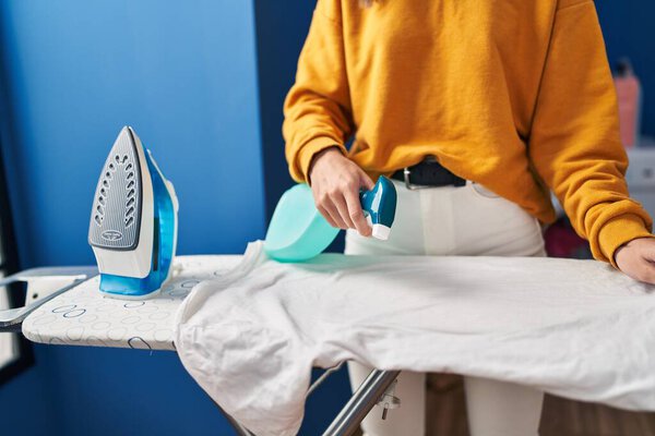 Young blonde woman ironing clothes applying liquid on t shirt at laundry room