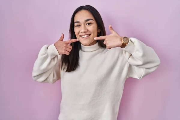 Young south asian woman standing over pink background smiling cheerful showing and pointing with fingers teeth and mouth. dental health concept.