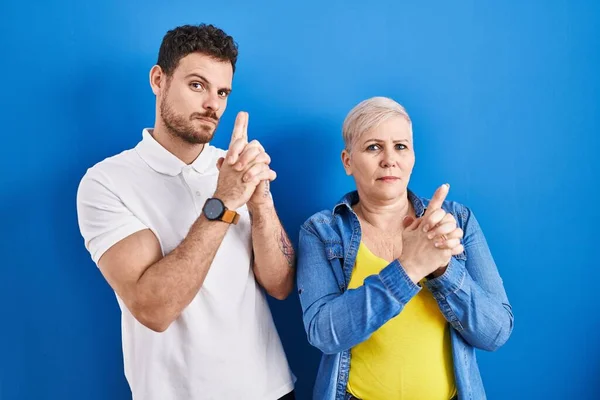 Young brazilian mother and son standing over blue background holding symbolic gun with hand gesture, playing killing shooting weapons, angry face
