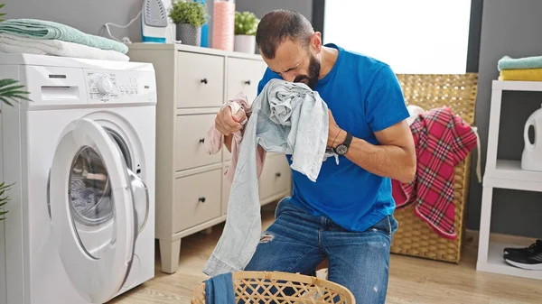 Young hispanic man washing clothes smelling dirty clothes at laundry room
