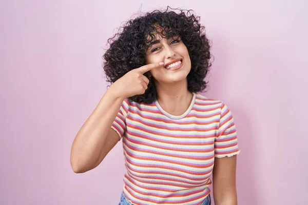 Young middle east woman standing over pink background pointing with hand finger to face and nose, smiling cheerful. beauty concept