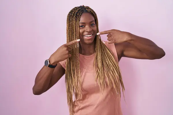 身穿辫子的非裔美国妇女站在粉红的背景上 笑容满面 用手指和嘴指尖 牙齿健康概念 — 图库照片