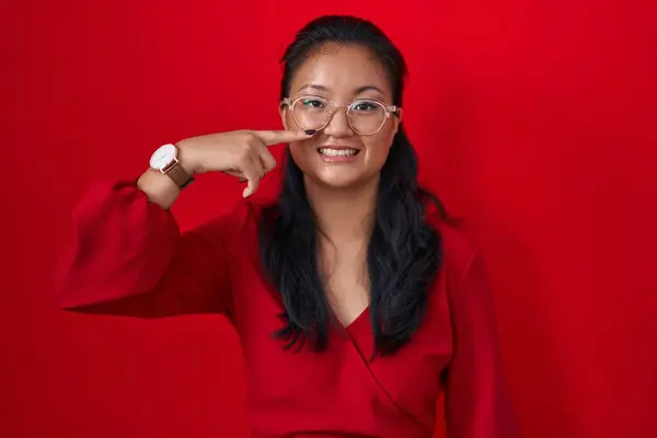 アジアの若い女性は 顔と鼻を指して赤い背景の上に立って 陽気で微笑んでいます 美容コンセプト — ストック写真